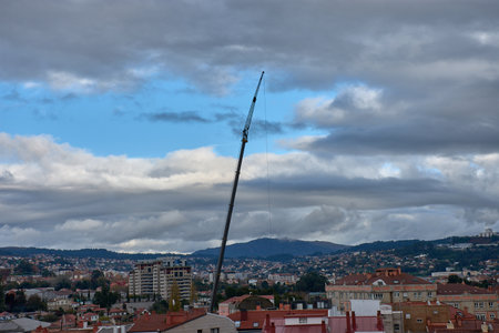 A tall crane rises above a residential area with red rooftops, framed by hills and a densely cloudy sky. The scene conveys a contrast between industrial activity and everyday life, evoking urban transformation, progress, and atmospheric tension. Ideal for editorial projects on architecture, urban planning, infrastructure, and the evolution of cities.の写真素材