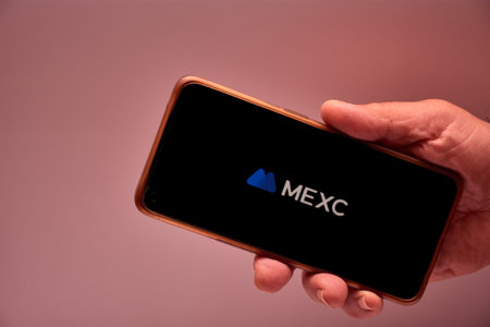 Vigo, Spain, November 30, 2025.A human hand holds a smartphone whose screen displays the MEXC logo, a cryptocurrency exchange platform renowned for its mega-trade matching technology. The blue background and clean design reinforce the technological and coのeditorial素材