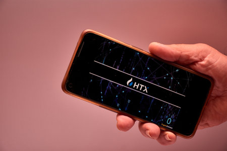 Vigo, Spain, November 30, 2025.A human hand holds a smartphone whose screen displays the logo of Huobi Global, an international digital asset platform founded in China in 2013. The dark background and clean design reinforce the technological and corporateのeditorial素材