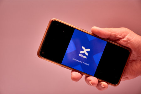 Vigo, Spain, November 30, 2025.A human hand holds a smartphone whose screen displays the BingX logo, an international cryptocurrency exchange platform with over 10 million users. The dark background and minimalist composition reinforce the technological aのeditorial素材