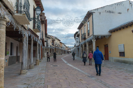 Ampudia, Palencia, Spain, December 7, 2025,A cobbled street in Ampudia stretches between wooden arcades and stone columns, framed by traditional Castilian architecture. The gray sky and festive lights create a cozy winter atmosphere, while several people のeditorial素材