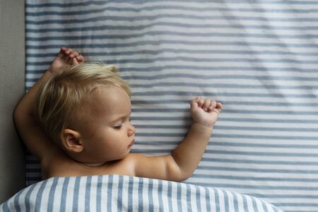 One year cute blond baby sleeps, raising hands up, on white and blue sheets. Light photo, good backgroundの写真素材