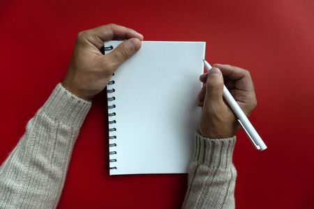 Mans right hand holds white pen and left hand holds a white papered blank notebook, view from the top, red background.の写真素材