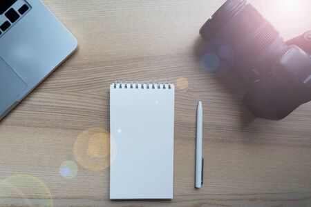 Photographer lay camera on wooden table near white paper notebook, white pen and modern laptop. Closeup of working place of creative person in sunny morningの写真素材
