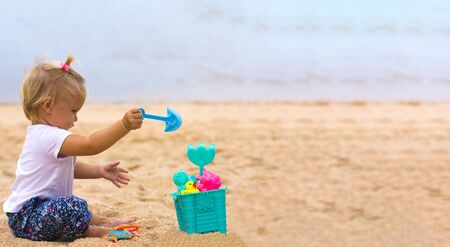 Cute little baby sits on sand on tropical beach and plays with toys.の写真素材