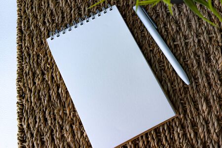 White papered spiraled notebook, white pen and green plant at rattan rug on white background, view from the top. Good for new year motivation and planningの写真素材
