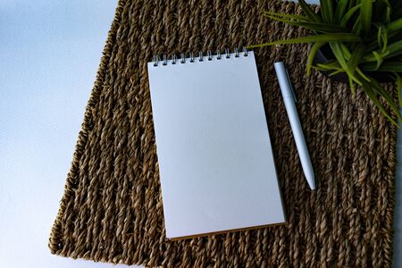 White papered spiraled notebook, white pen and green plant at rattan rug on white background, view from the top. Good for new year motivation and planningの写真素材