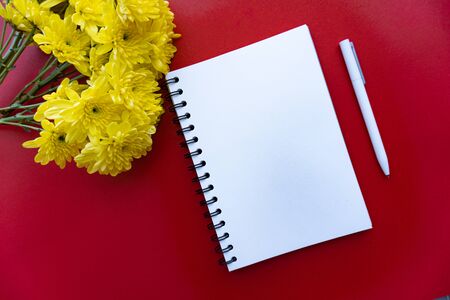 White papered spiraled notebook, white pen and yellow flowers at red background, view from the top. Good for new year motivation and planningの写真素材