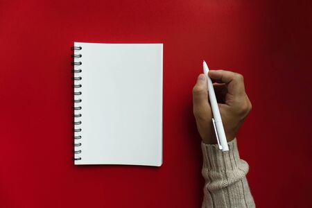 Mans right hand in winter sweater hold white pen and writes his plans and list of motivation for future for new year 2020 in white papered blank spiral notebook, red backfroundの写真素材