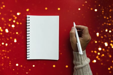 Mans in winter sweater holds pen and writes plans and list of motivation for future for new year 2020 in white papered blank spiral notebook, red backfround. Christmas and new year conceptの写真素材