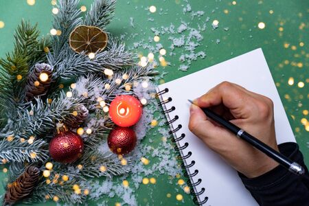 Man with black pen writes his goals for new year in blank notebook There are branches, xmas balls, red candle and snow on green background.の写真素材