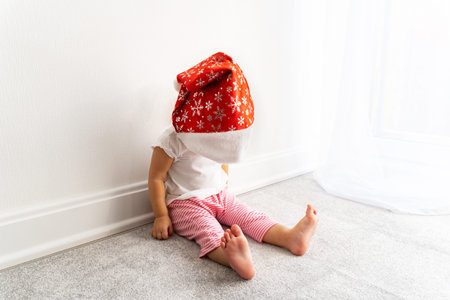 Baby in red santa claus hat sits on grey carpet near white wall and waits for christmas. New year concept. Funny kid, tired toddler.の写真素材