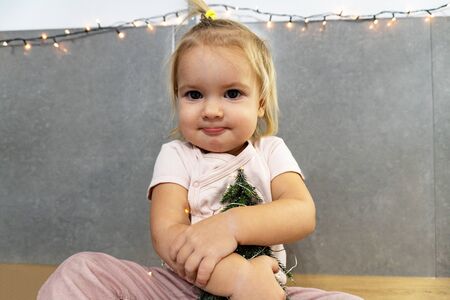 Cute blond happy baby girl hugs a green tree on grey stone background with Christmas lights on the wall, new year conceptの写真素材