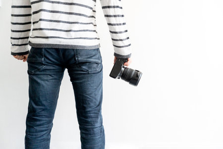 Man stands near white wall with digital camera in a hand. Man in black jeans and white striped...