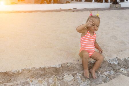 Cute baby girl in pink swimming suit sits on sand at the beach, raise hand and tries to close from the bright sun.の写真素材