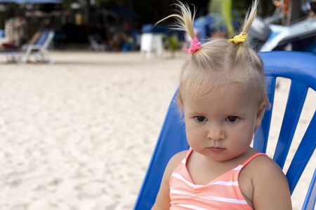 Portrait of serious cute baby in pink swimming suit sitting on plastic chair at the sand at the sea at summer asian resort.の写真素材