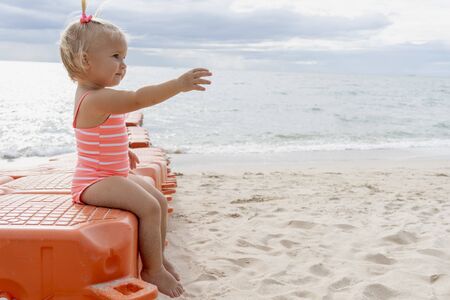 Cute blond baby in pink swimming suit sits on modular floating pontoon near sea at asian resort and shows something with her right hand. Summer sunny day.の写真素材