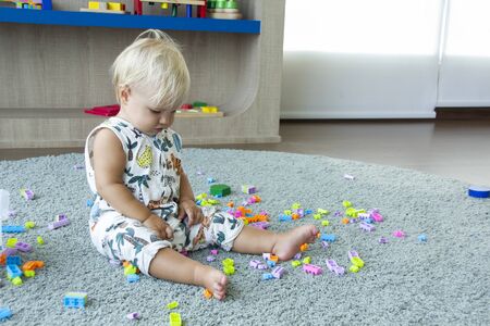 Small cute blond baby sits on a rug and plays with colourful toy bricks around. Toddler in a kinder room. Good for bannersの写真素材