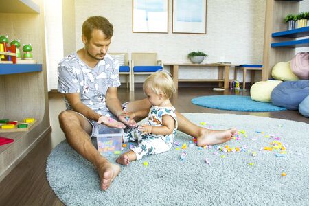 Father plays with his cute blond baby in kindergarten on big grey rug with bricks. Sun shining through the window. Children and father concept. Good background. Space for the text.の写真素材