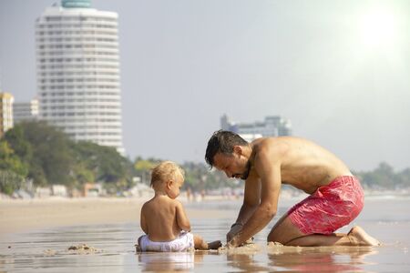 Father and little caucasian blondy cute kid playing with sand at the beach near the sea in summer. Happy family time. Sunny day.の写真素材