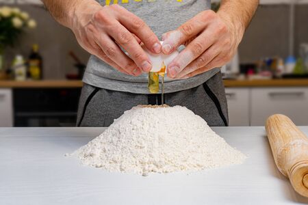 Man at kitchen. His hands tries to open or break egg above white gluten flour. Close up of process of preparing dough for baking.の写真素材