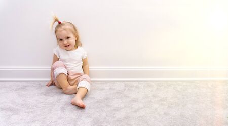 Cute bare foot baby girl in pink trousers sits near white wall on grey rug, smiles and closes eyes from sun, good background, copy spaceの写真素材