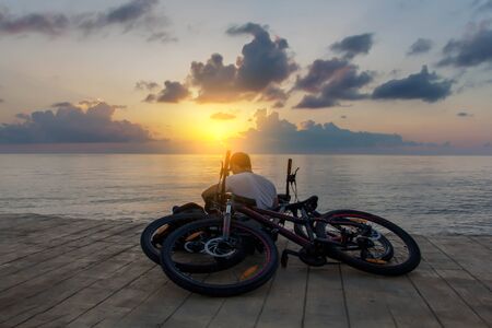 After cycling man in white short sits near sea and works or dreams or writes. View from the back. His bicycles near him. Sunset or sunrise.の写真素材