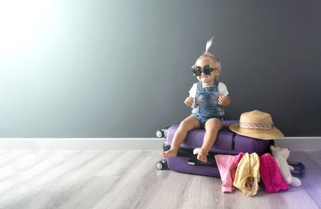 Little cute barefoot blond baby girls in sunglasses sits on a violet suitcase and waits for summer holiday travel trip in abroadの写真素材