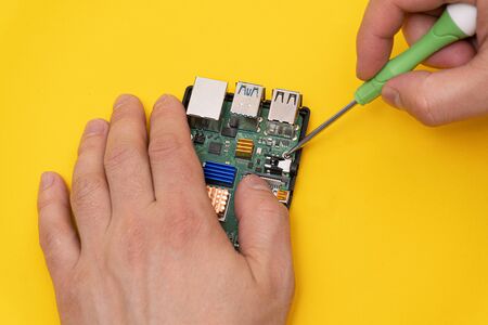 Man fixes circuit board in microcomputer with screwdriver with green handle on yellow background, closeup.の写真素材