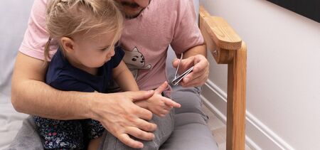 Father cuts child nails to his baby with clipper. The kid sits at male knees. Manicure at homeの写真素材