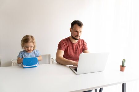 Young man, father should work from home during quarantine time cause of coronavirus. Child at home, kindergarden closed. Baby helps father to work. They work in laptops. White background.の写真素材