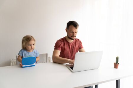 Young man, father should work from home during quarantine time cause of coronavirus. Child at home, kindergarden closed. Baby helps father to work. They work in laptops. White background.の写真素材