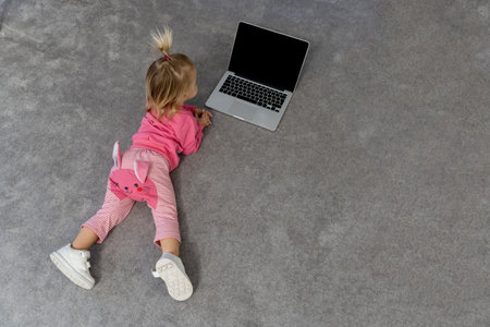 Small cute blond caucasian interested baby laying on grey rug on floor and watching tv on laptop,...