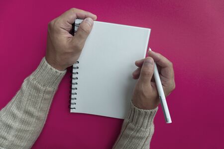 Mans right hand in winter sweater hold white pen and writes his plans and list of motivation for future for new year in white papered blank spiral notebook, pink backfroundの写真素材