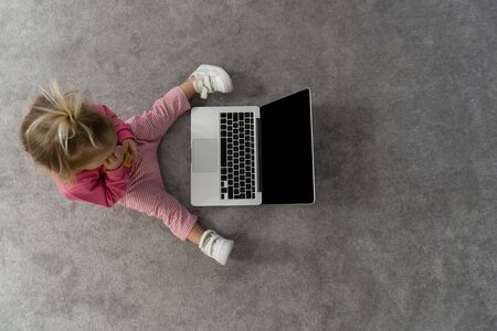 Small cute blond caucasian baby laying on floor on grey rug and watching cartoons on laptop with great interest. black screen, mockup, copy space,view from the topの写真素材