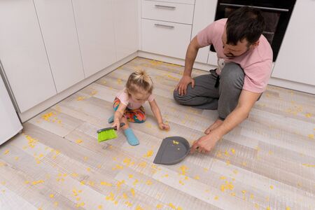 Cute little girl helps her father with cleaning the floor from cornflakes at the kitchen. Concept of family housework. Copy space.の写真素材