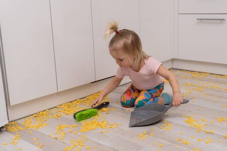 Cute little caucasian girl cleans the floor from cornflakes with scoop and brush at the kitchen. Copy space.の写真素材