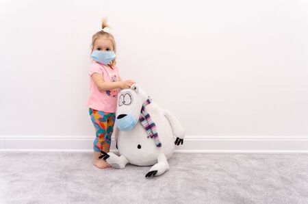 Little blond caucasian girl baby in protective mask sits isolated at home at quarantine with white bear toy in protective mask, to protect from spread of contagious covid-19 infectionの写真素材