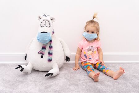 Little blond caucasian girl baby in facial protective mask sits at home at quarantine near bear toy in facial protective mask, precautions against coronavirus infection concept, white background.の写真素材