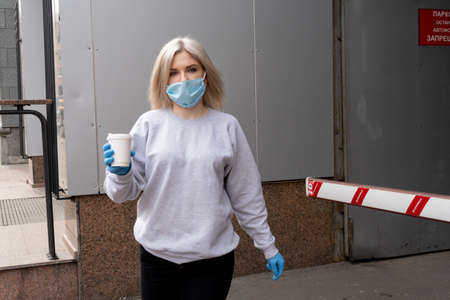 Kiev, Ukraine, March 27, 2020, Portrain of blond ukrainian woman in facial protective medical mask and mrdical gloves carries takeaway paper coffee cup.のeditorial素材