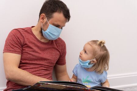Cute small girl in facial medical mask looks at father who is in facial medical mask and reading book at family quarantine time, protection measures against coronavirus.の写真素材