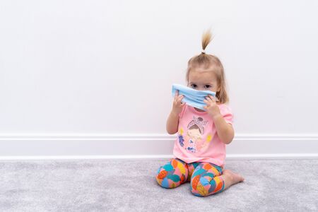 Small cute lovely child sits on floor at home at quarantine and carries facial protective medical mask. necessary protective measures against dangerous coronavirus disease. stay home concept.の写真素材