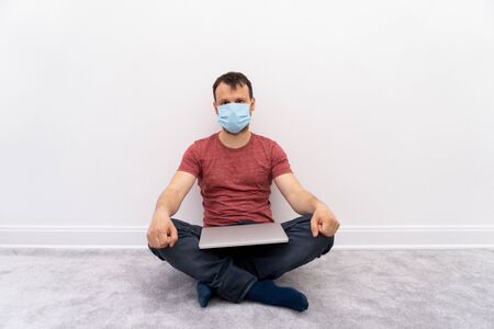 Man at self quarantine works from home in laptop at remote job, guy at protective surgical mask sits on floor. Protective measures from coronavirus.の写真素材
