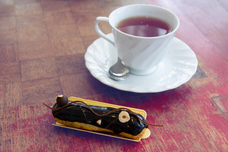 Closeup at white cup with black tea and eclair with chocolate on rustic table.
