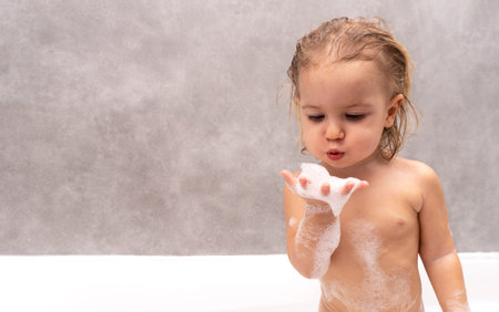 Cute caucasian naked kid plays with white foam at bathroom, happy childhood concept, grey...