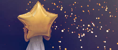Caucasian little child holding star balloon hiding face, black background with bokeh lights, christmas concept, birthday celebration, toned.の写真素材