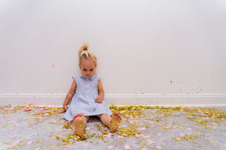 Cute caucasuian blond baby girl sits on a rug with a lot of gold tinsels after celebration holiday,...