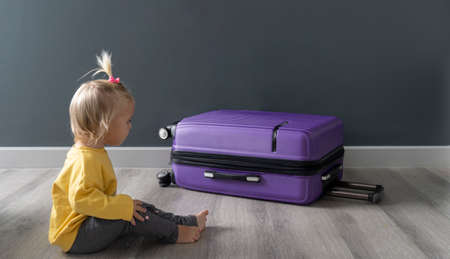 Little happy cute blond kid sitting on floor and looking at violet luggage, ready for interesting journey after quarantine, lockdown, due to covid. Family travel concept. Copyspace.の写真素材
