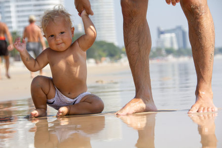 Cute blond caucasian baby is learning to foot with parents hand at the sandy beach, summer time.