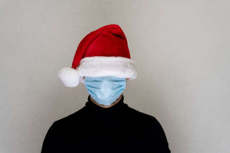 Caucasian man with hidden face under blue facial medical protective mask and red santa hat, new reality due to coronavirus, copy space.の写真素材